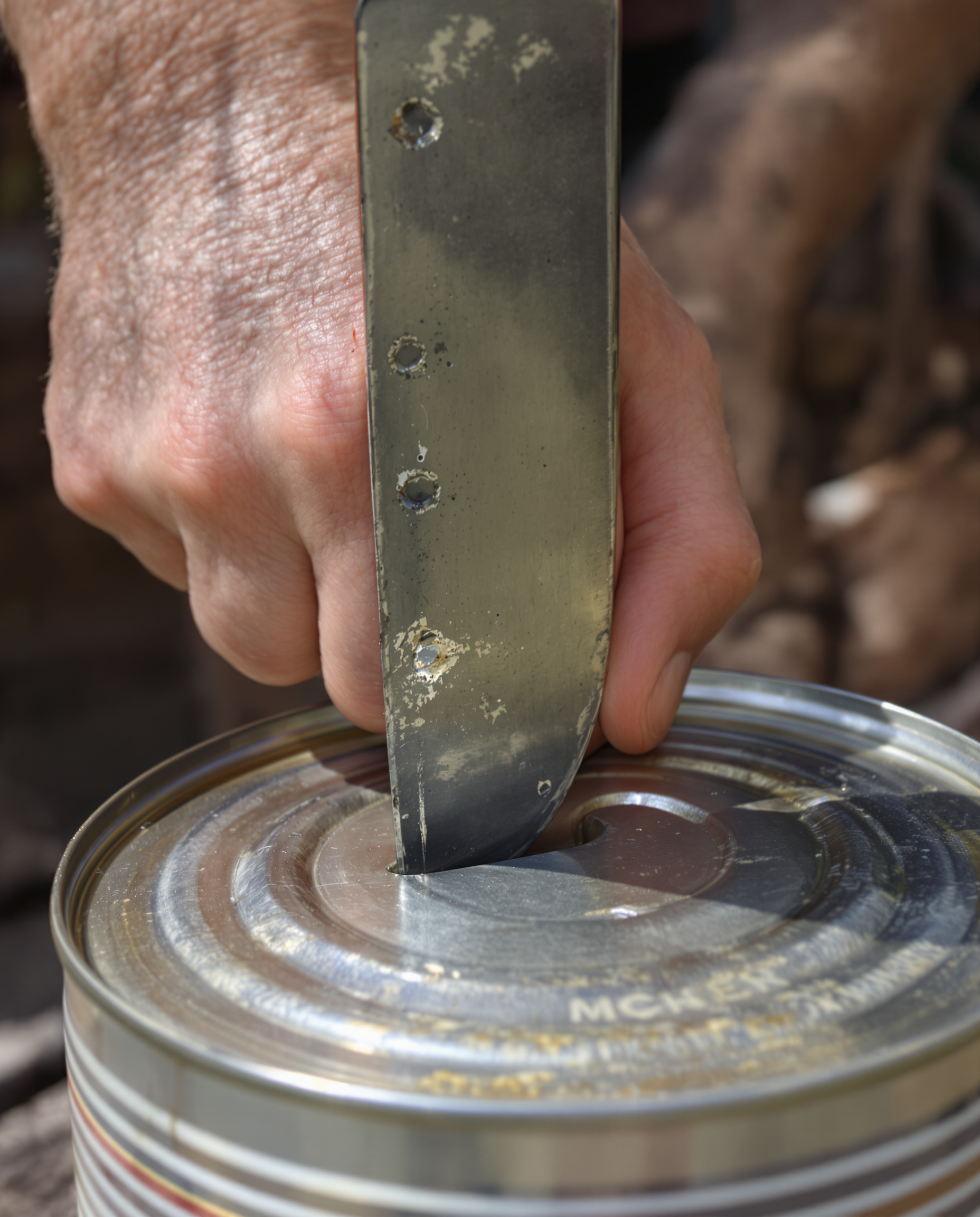 Gros plan sur une main ferme tenant un couteau de chef à lame fixe, la pointe appuyée verticalement sur le couvercle d'une boîte de conserve. On devine plusieurs marques de poinçonnement déjà réalisées le long du rebord.