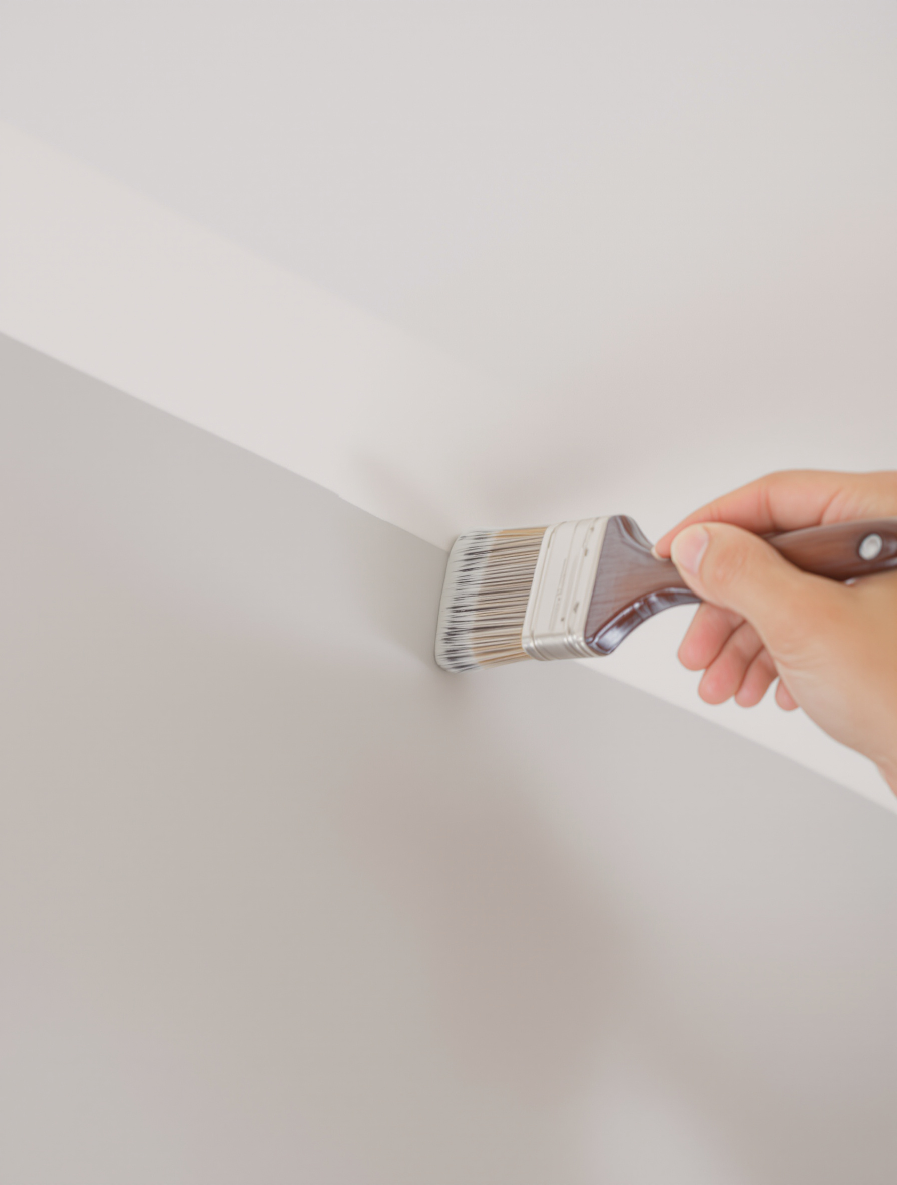 Gros plan sur une main experte utilisant un pinceau à réchampir pour peindre avec précision l'angle entre un mur gris clair et un plafond blanc. La lumière met en valeur la netteté de la ligne.