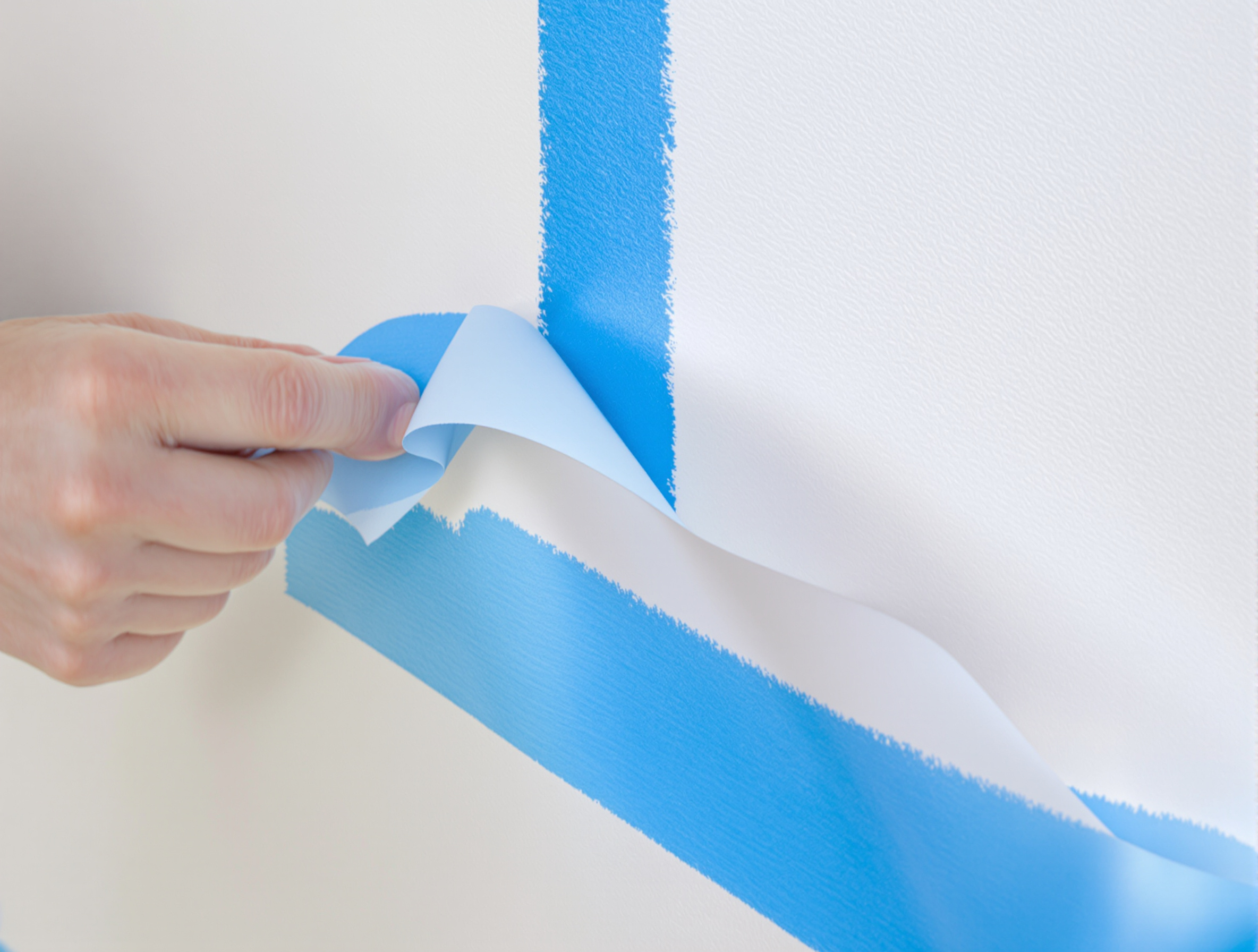 Retrait délicat d'un ruban de masquage bleu sur un mur fraîchement peint, angle visible à 45°. La démarcation est nette.